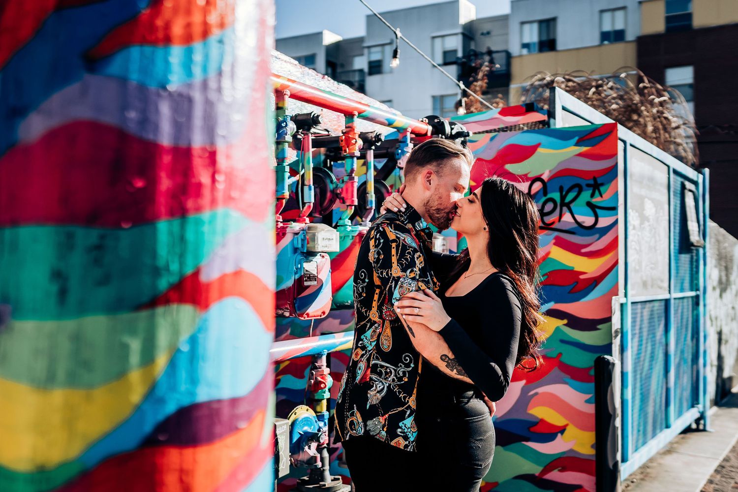 Couple embraces in front of colorful graffiti wall with vibrant rainbow swirls and patterns.
