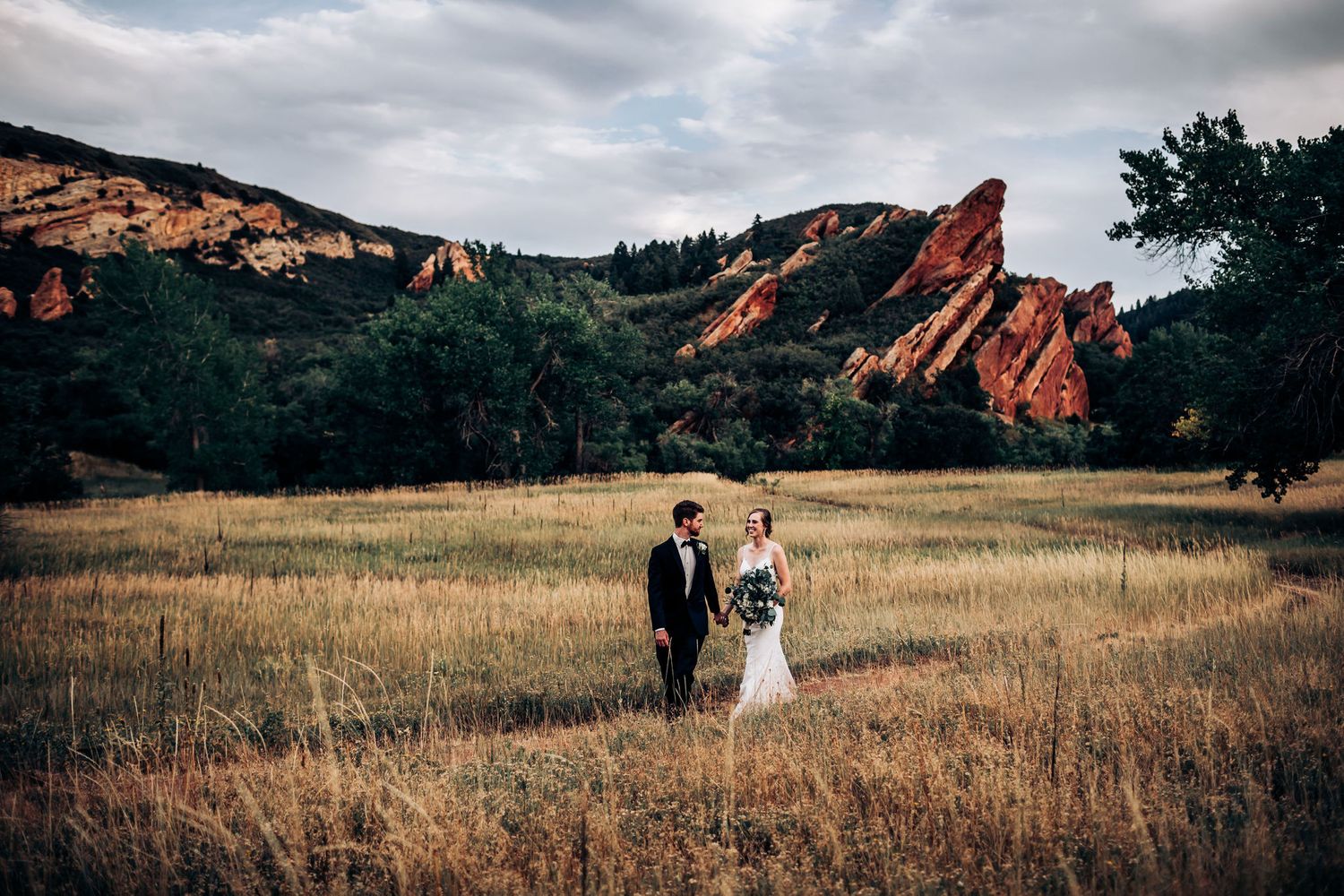 Wedding couple stands in golden field with striking red rock formations in background.