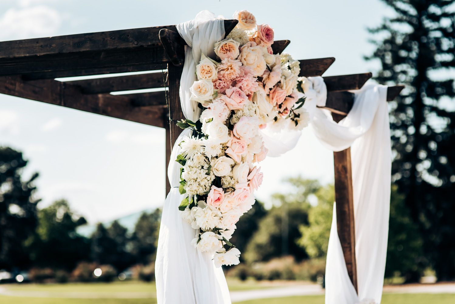 Wedding arch decorated with pink and white floral arrangements and flowing white fabric outdoors.