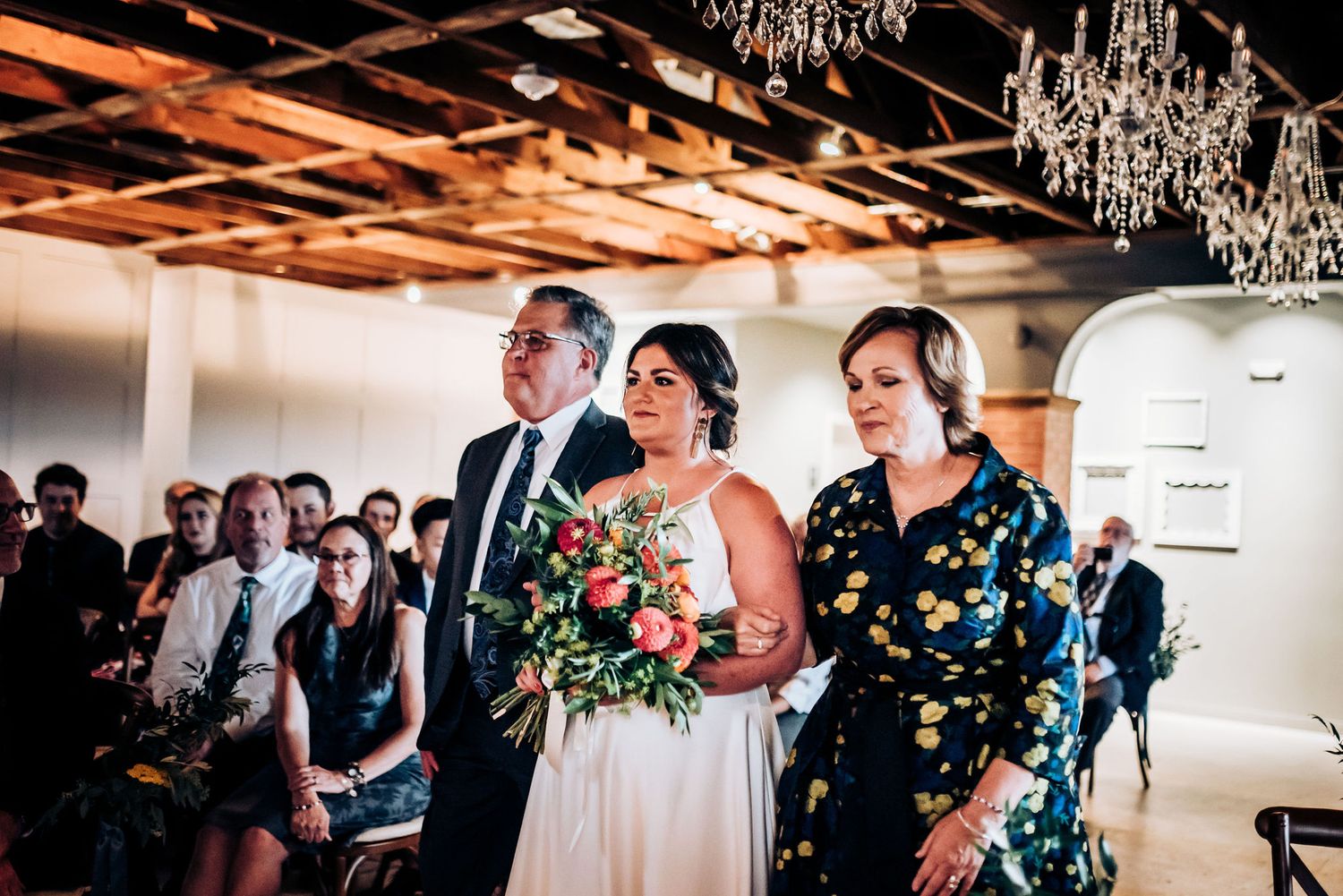 Wedding ceremony in a rustic indoor venue decorated with crystal chandeliers and exposed wooden beams.