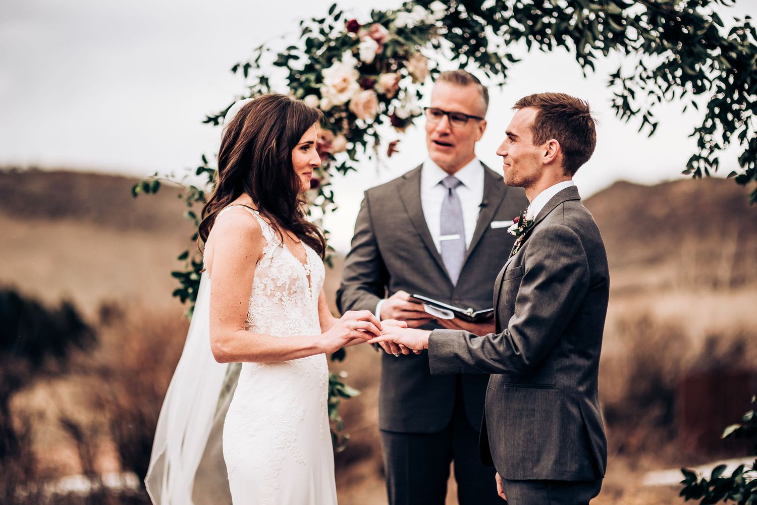 Wedding vow exchange beneath floral arch with rolling hills in background.