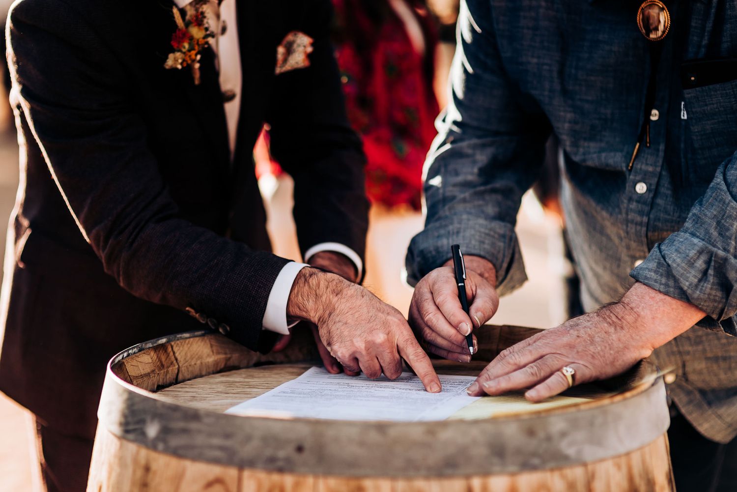 Hands signing documents on rustic wooden barrel during outdoor ceremony.