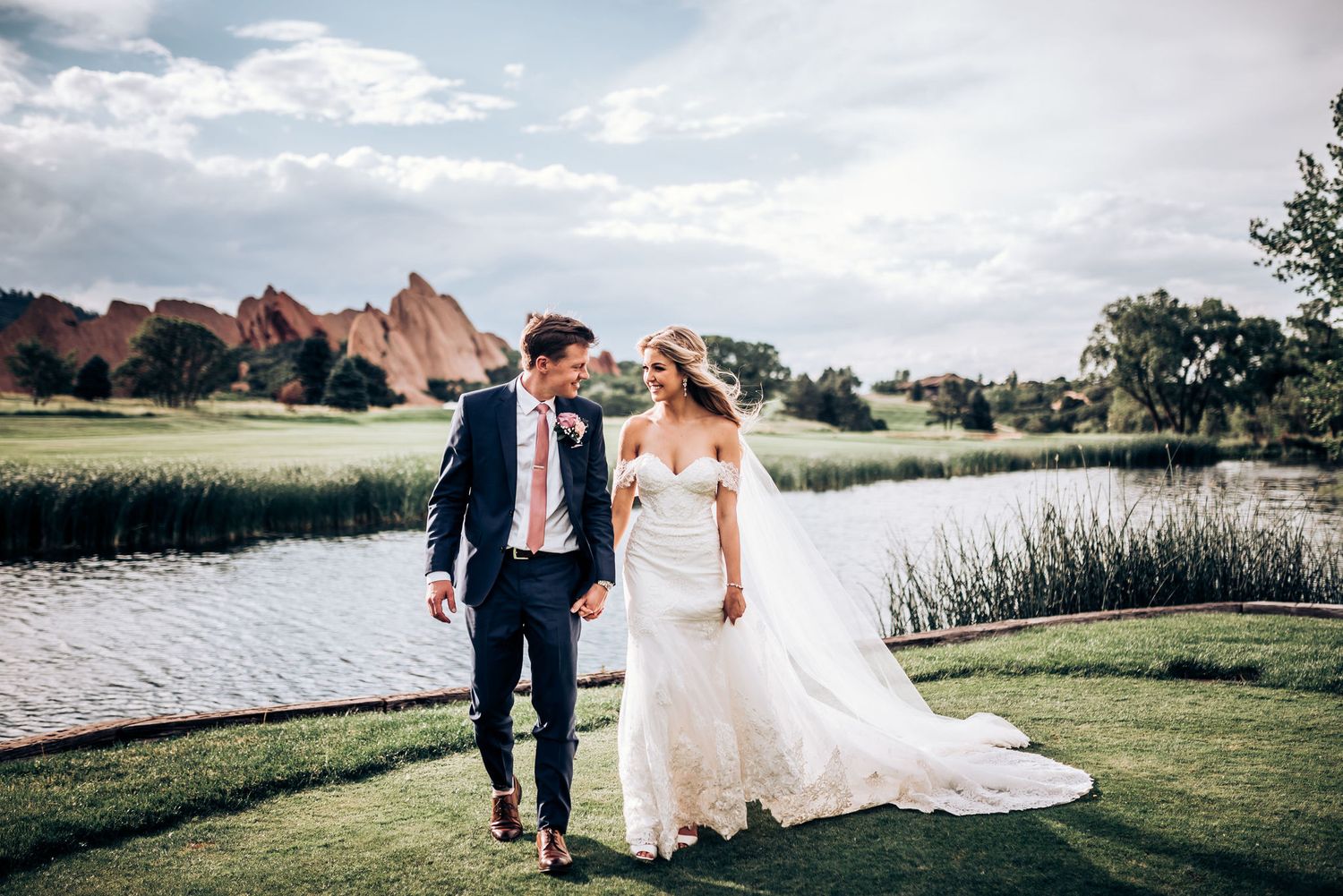 Wedding couple strolls along lakeside path with majestic red rock formations in background.