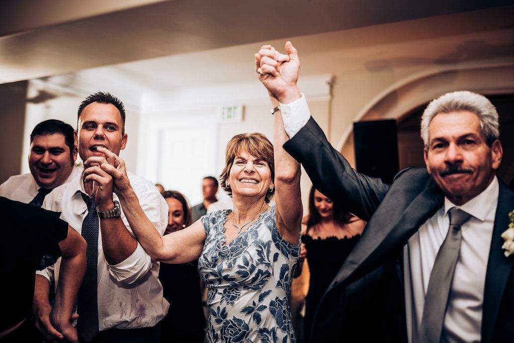 Wedding guests raise their hands in celebration during a lively reception party under arched ceilings.