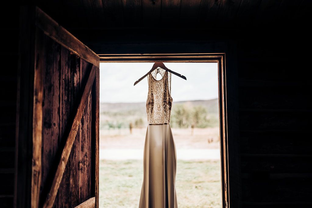 A sleeveless wedding gown silhouetted in a rustic barn doorway with mountain landscape.