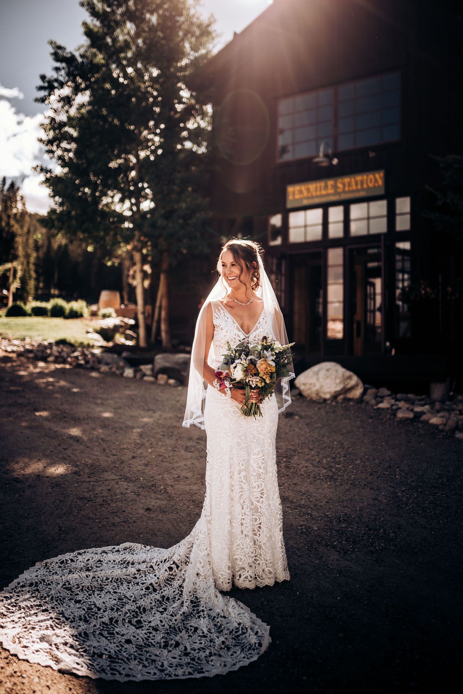 Sunlit bridal portrait in front of rustic building with dramatic train and wild bouquet.