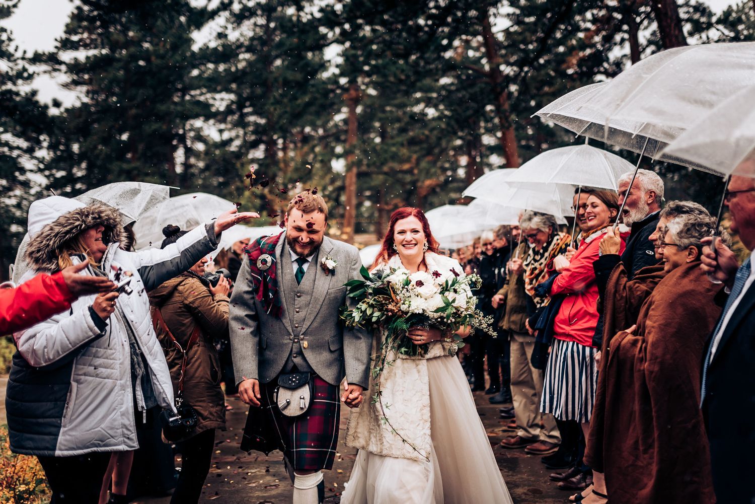 Wedding party walks through crowd of guests holding umbrellas in winter forest setting.