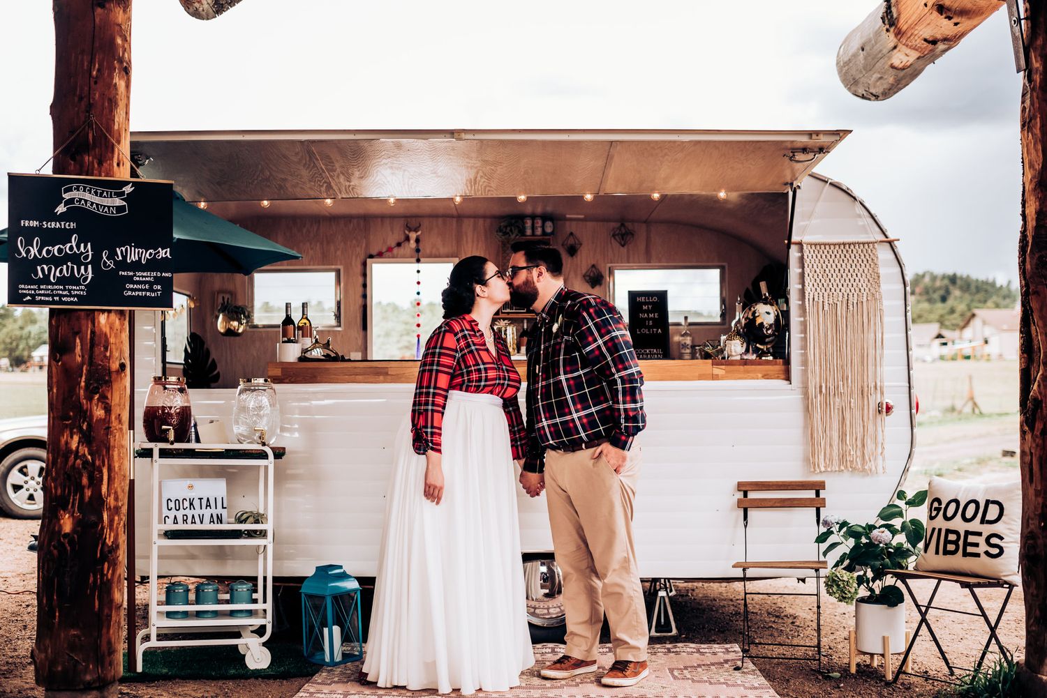 Stylish couple in plaid and white dress stand by vintage food truck in desert setting.