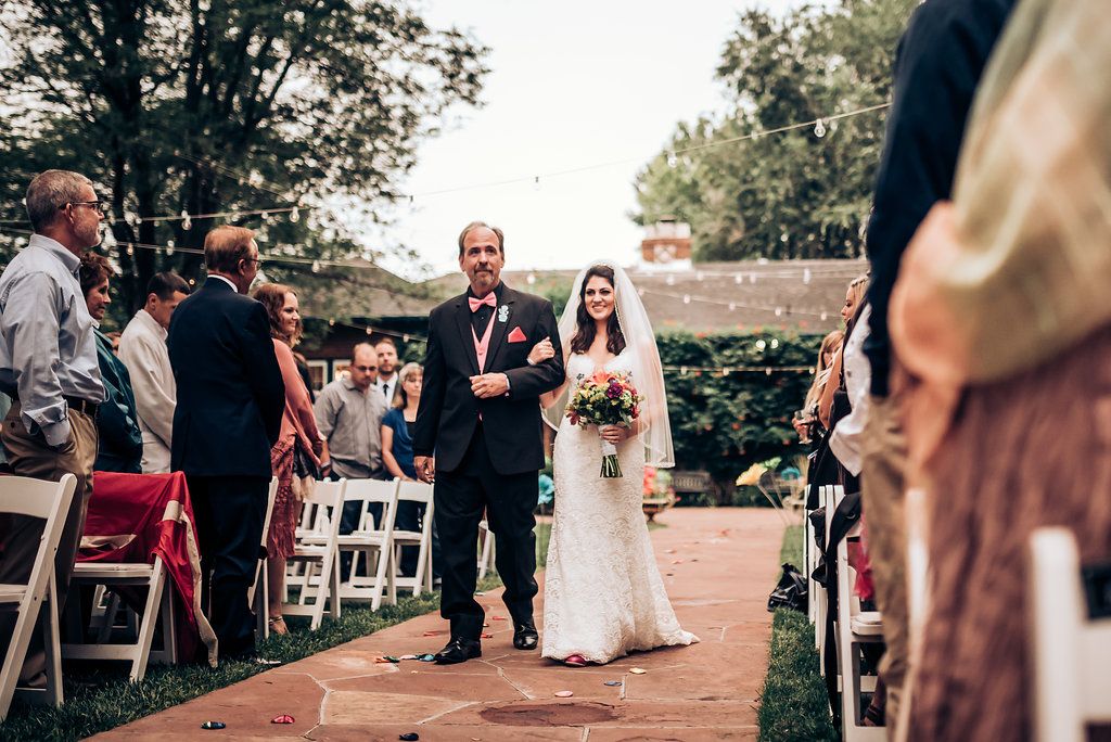 Wedding ceremony processional on a brick pathway with guests seated on white chairs outdoors.