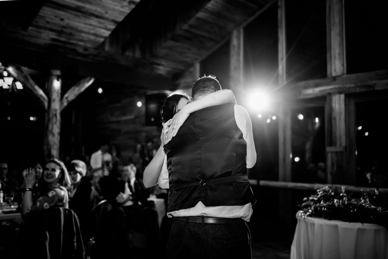 A black and white shot of a wedding reception dance in a rustic venue with backlighting.