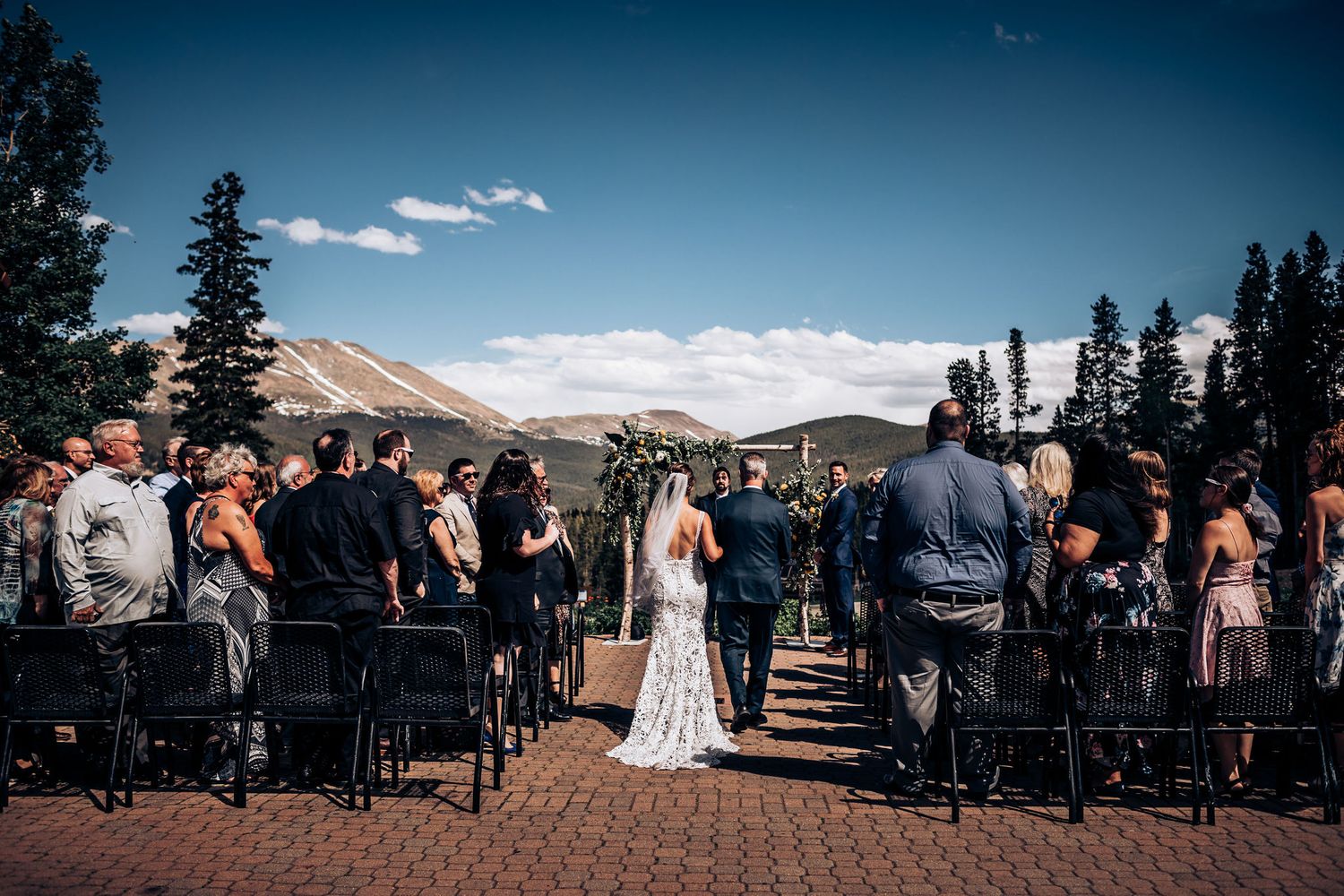 Wedding guests seated at an outdoor mountain venue watching a ceremony under blue skies.