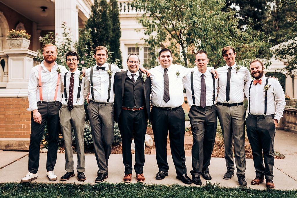 Wedding party members in coordinated attire with suspenders stand in formation on garden grounds.