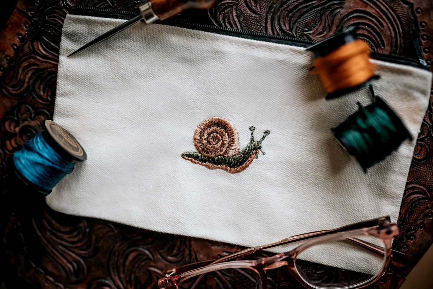 White canvas pouch with embroidered snail design surrounded by colorful thread spools.