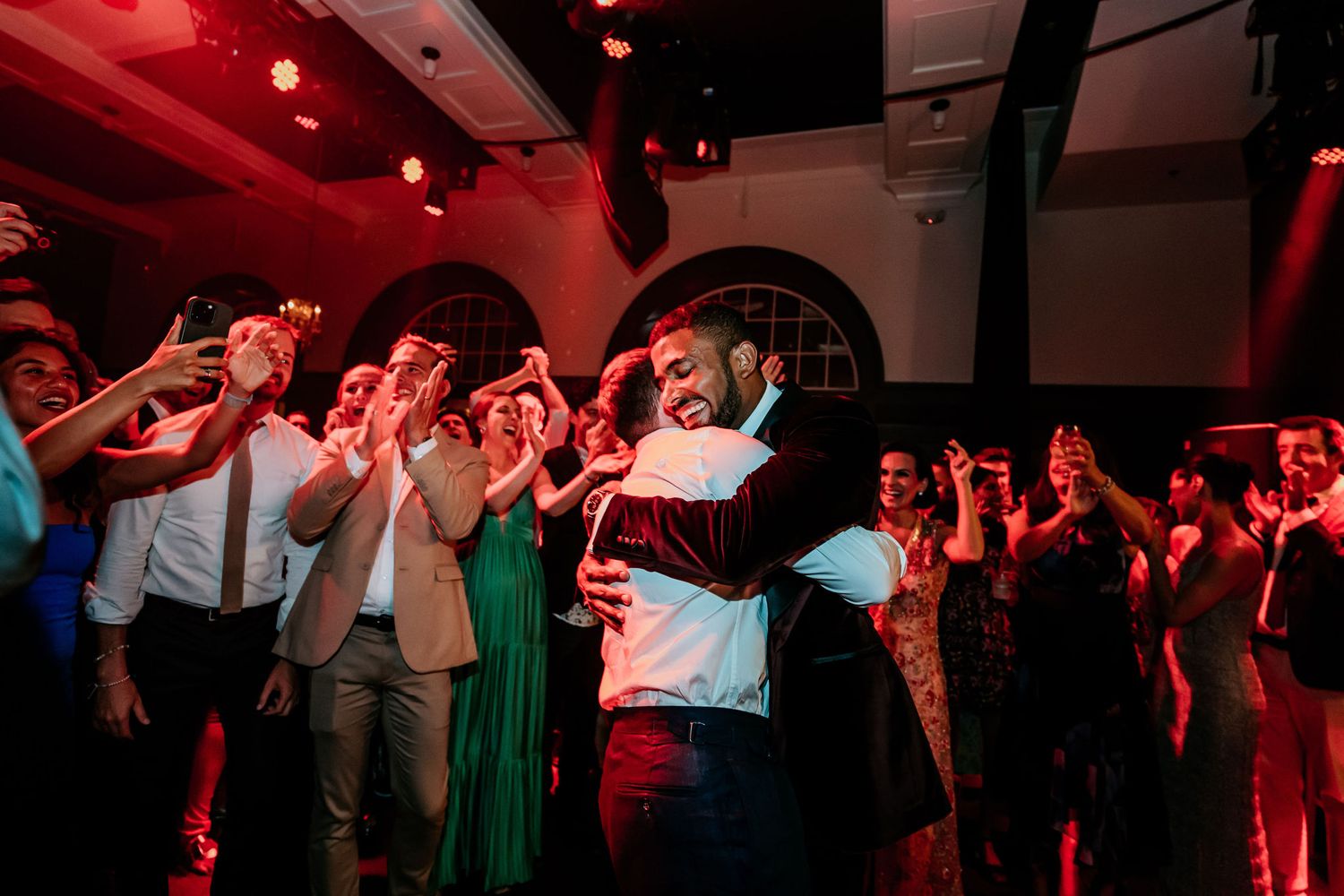 Lively wedding reception with guests dancing under red lighting and arched windows.