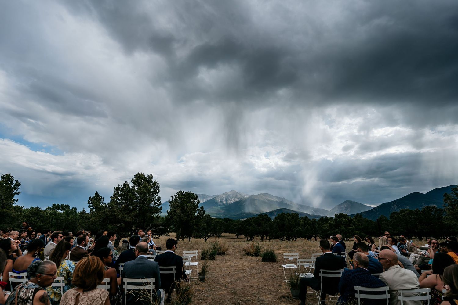 Wedding guests seated at outdoor ceremony with dramatic stormy mountain backdrop.