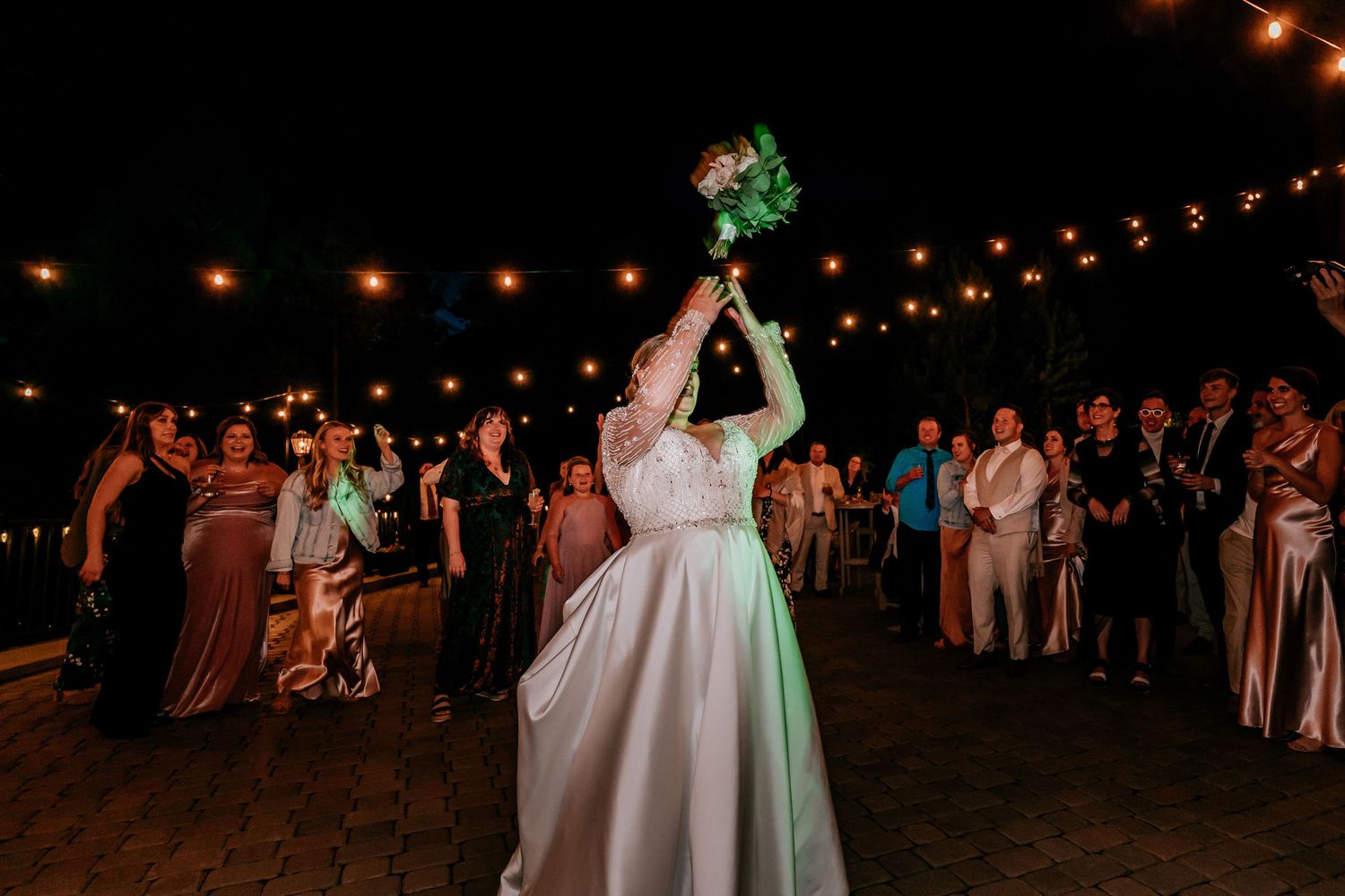 Bride tosses bouquet under string lights at nighttime wedding reception.