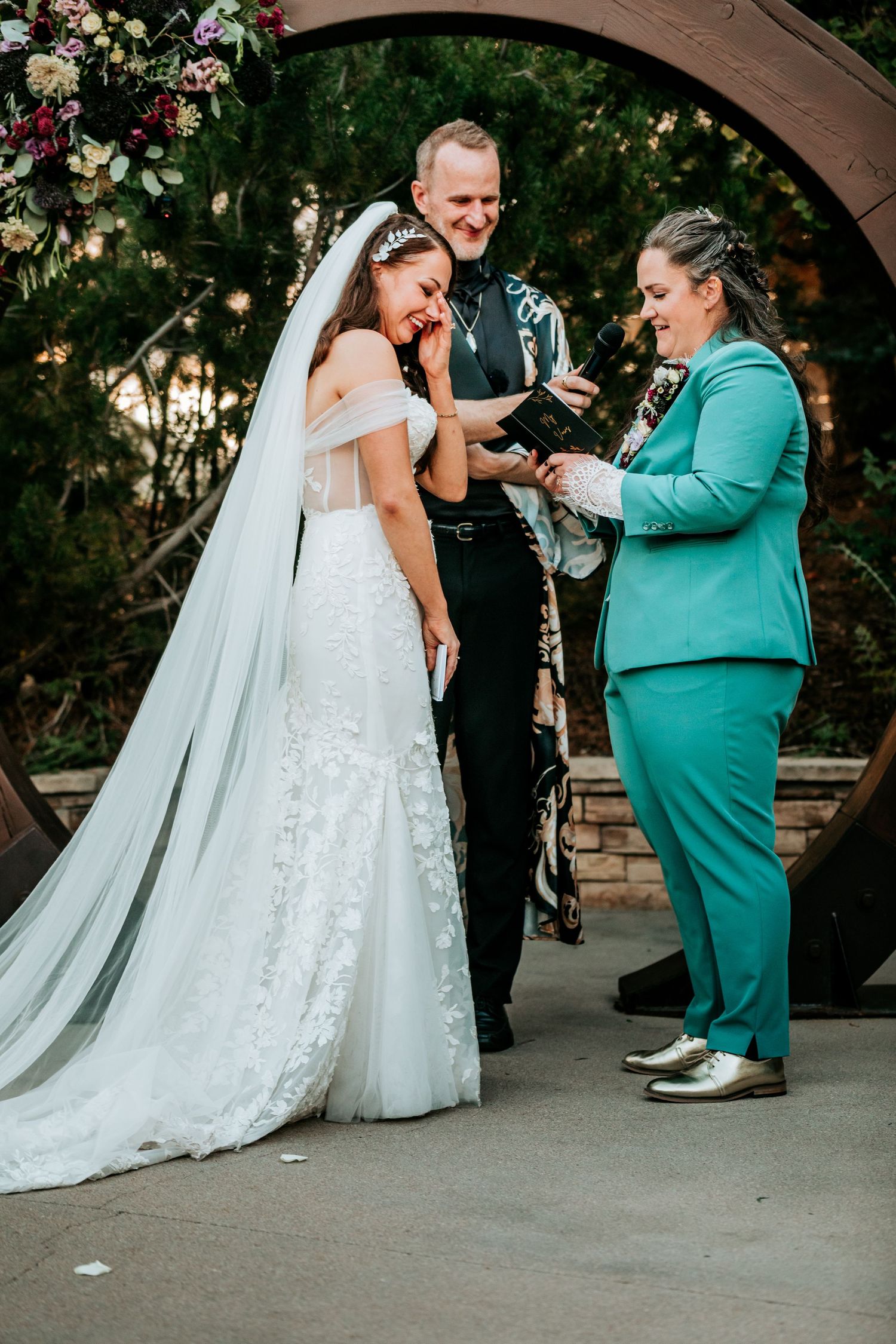 Bride laughing joyfully with partner in teal suit during ring exchange at outdoor wedding ceremony.