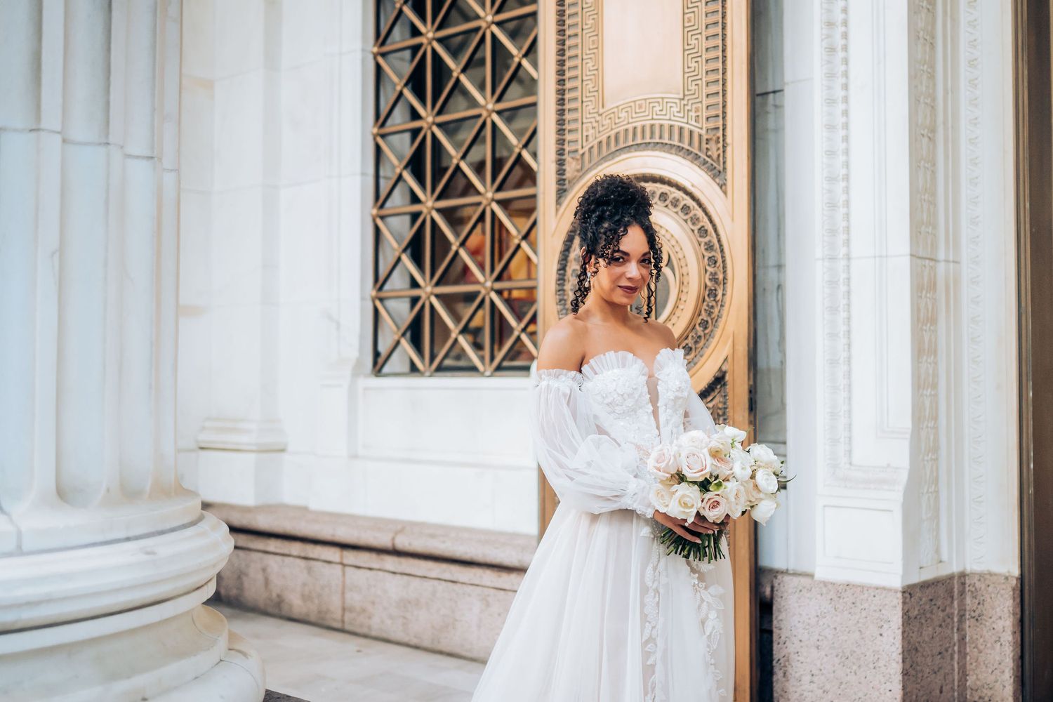 Bride in elegant white gown holds bouquet against architectural backdrop with ornate details.