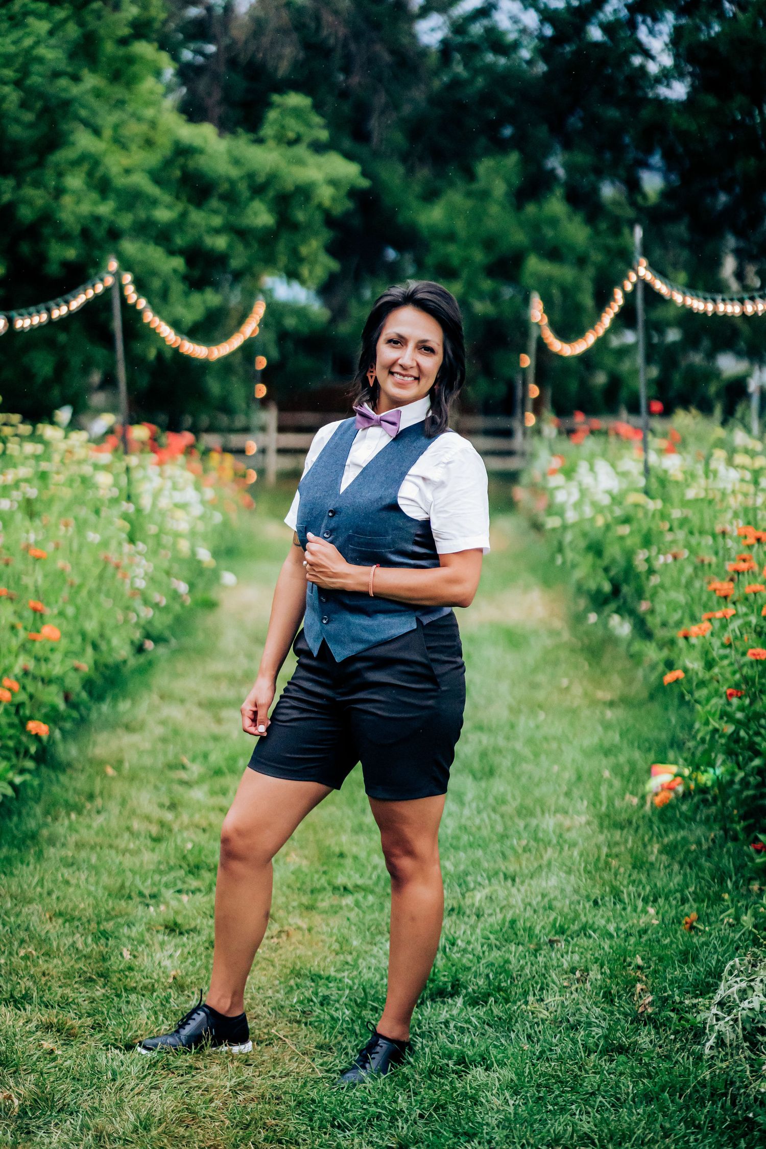 Person in formal vest and shorts smiling in a garden path with string lights overhead.