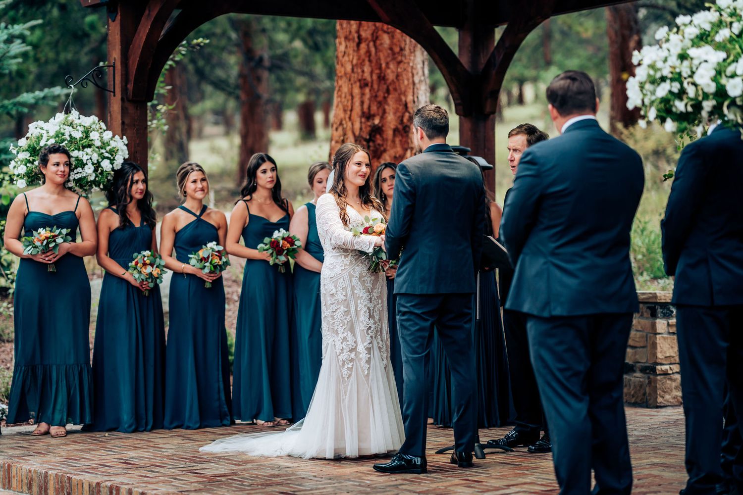 A bridal party in navy blue dresses and suits stands under a rustic wooden pavilion during the ceremony.