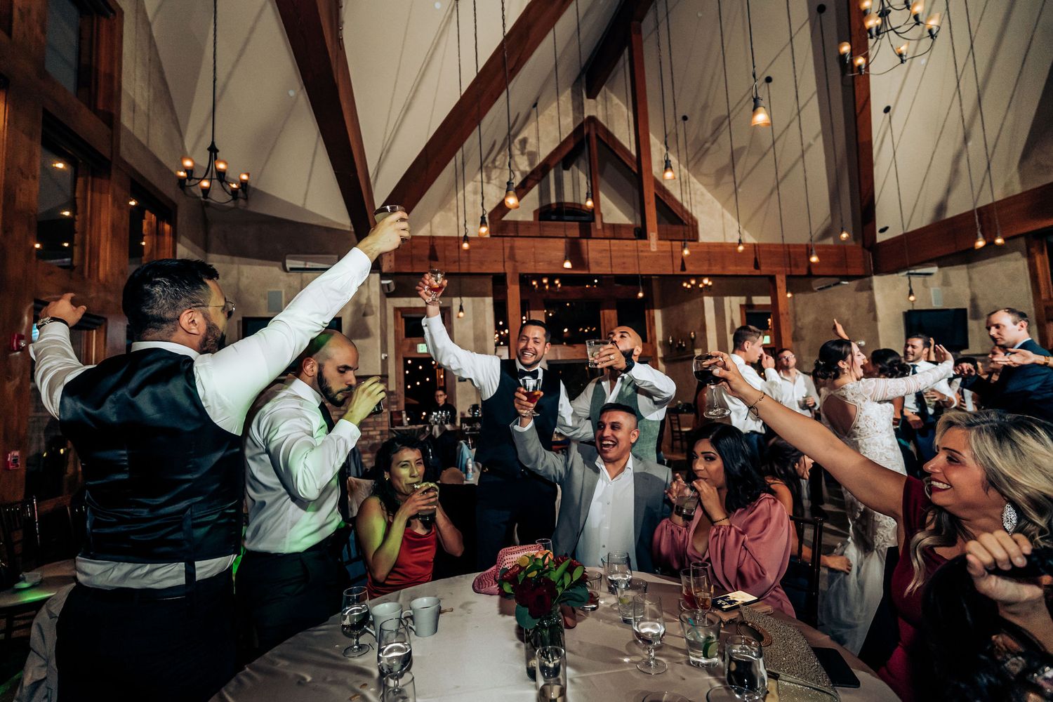 Wedding guests raise glasses in celebration during reception toast at rustic venue with wooden beams.