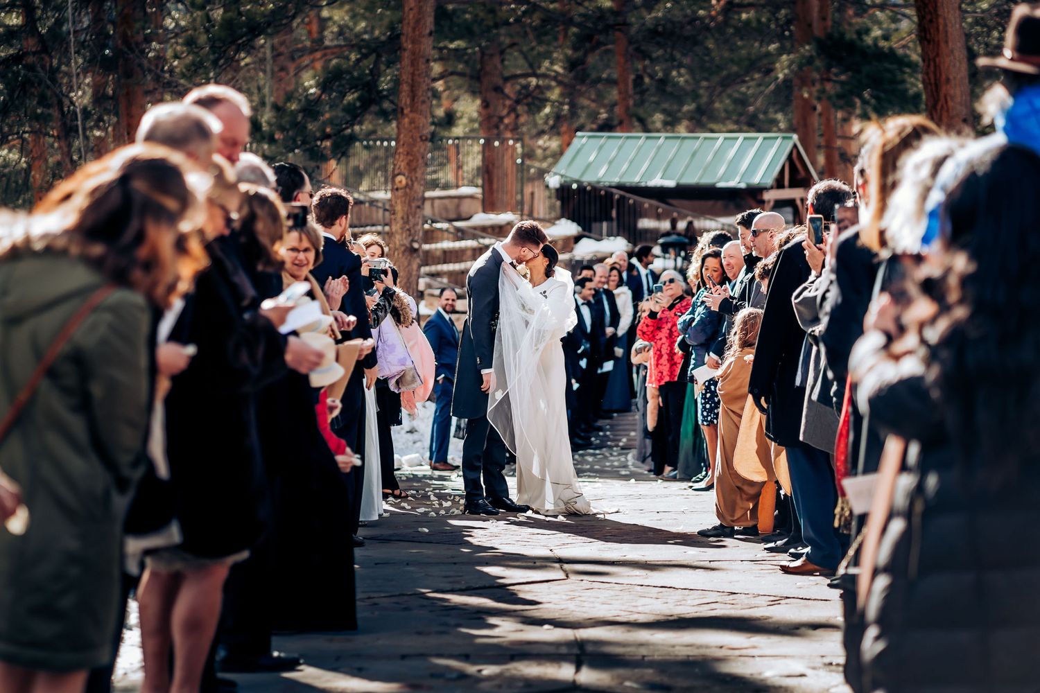 Wedding couple embraces while guests form aisle in snowy forest setting.