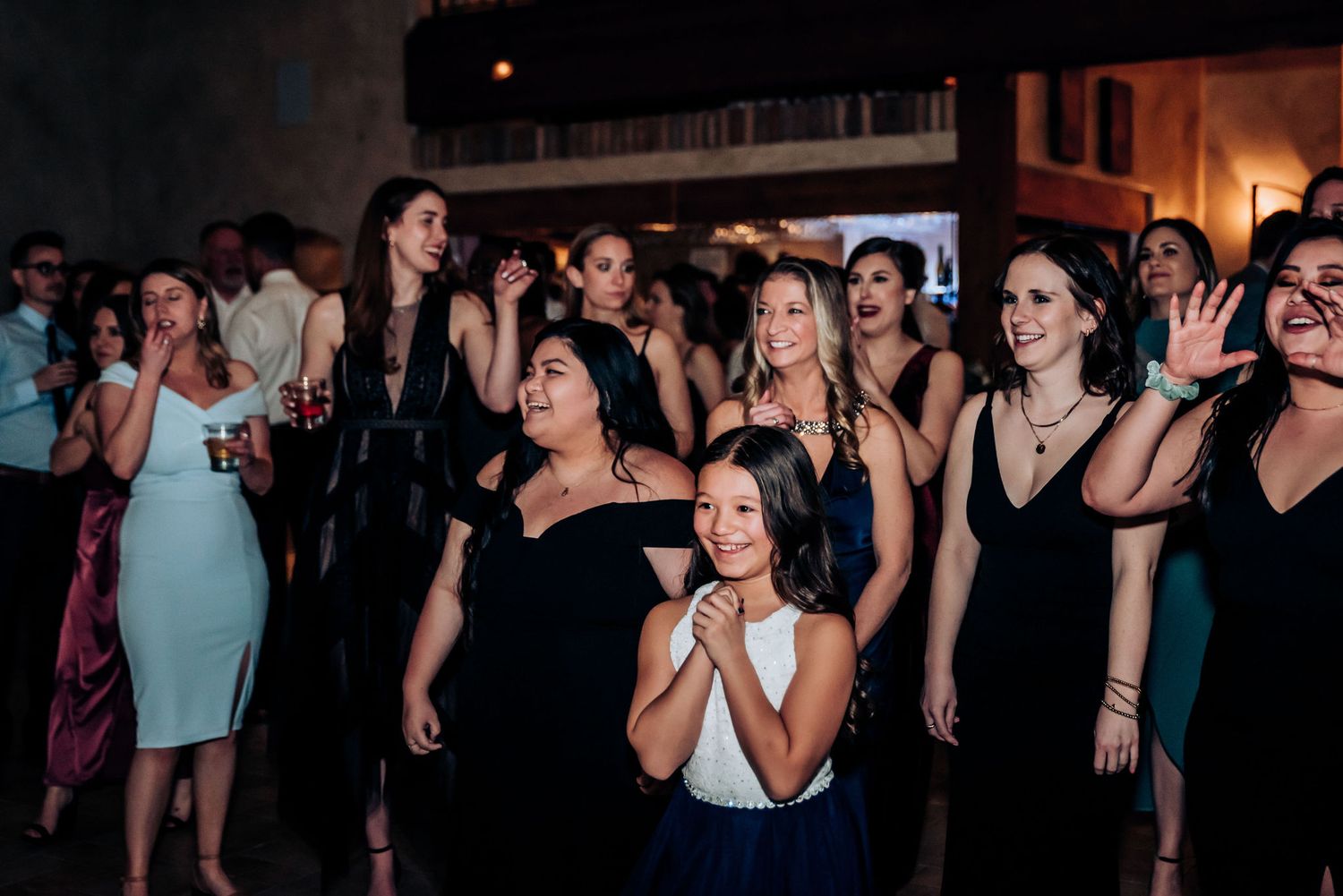 Group of wedding guests in dark dresses sing and dance together during the reception.