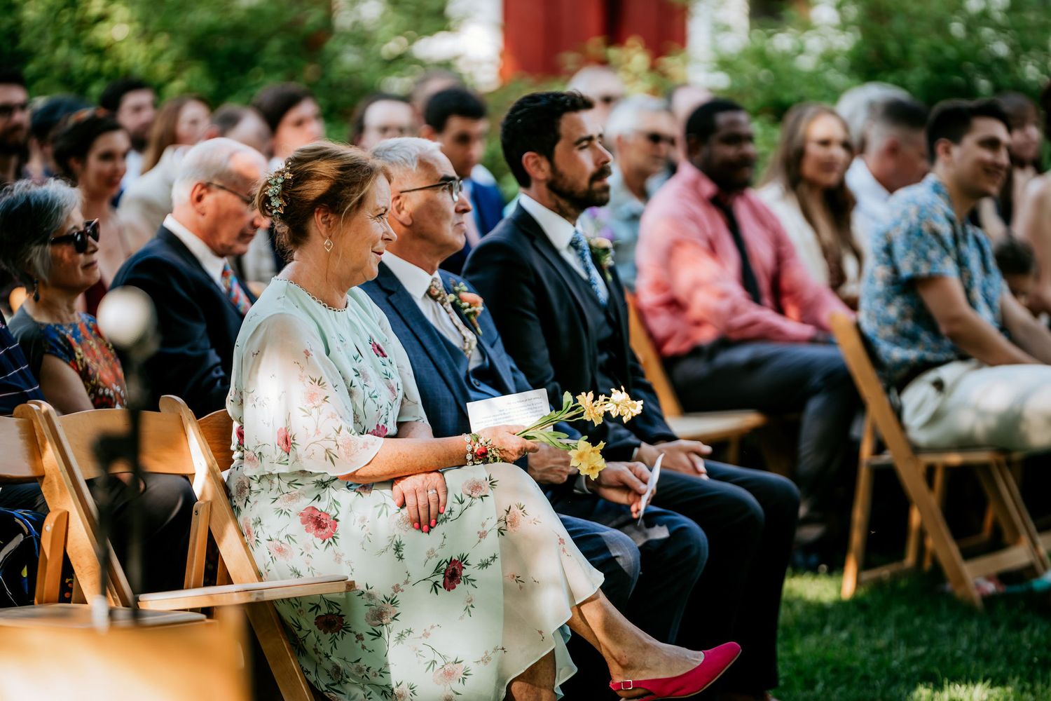 Wedding guests seated in wooden chairs outdoors watch a ceremony.