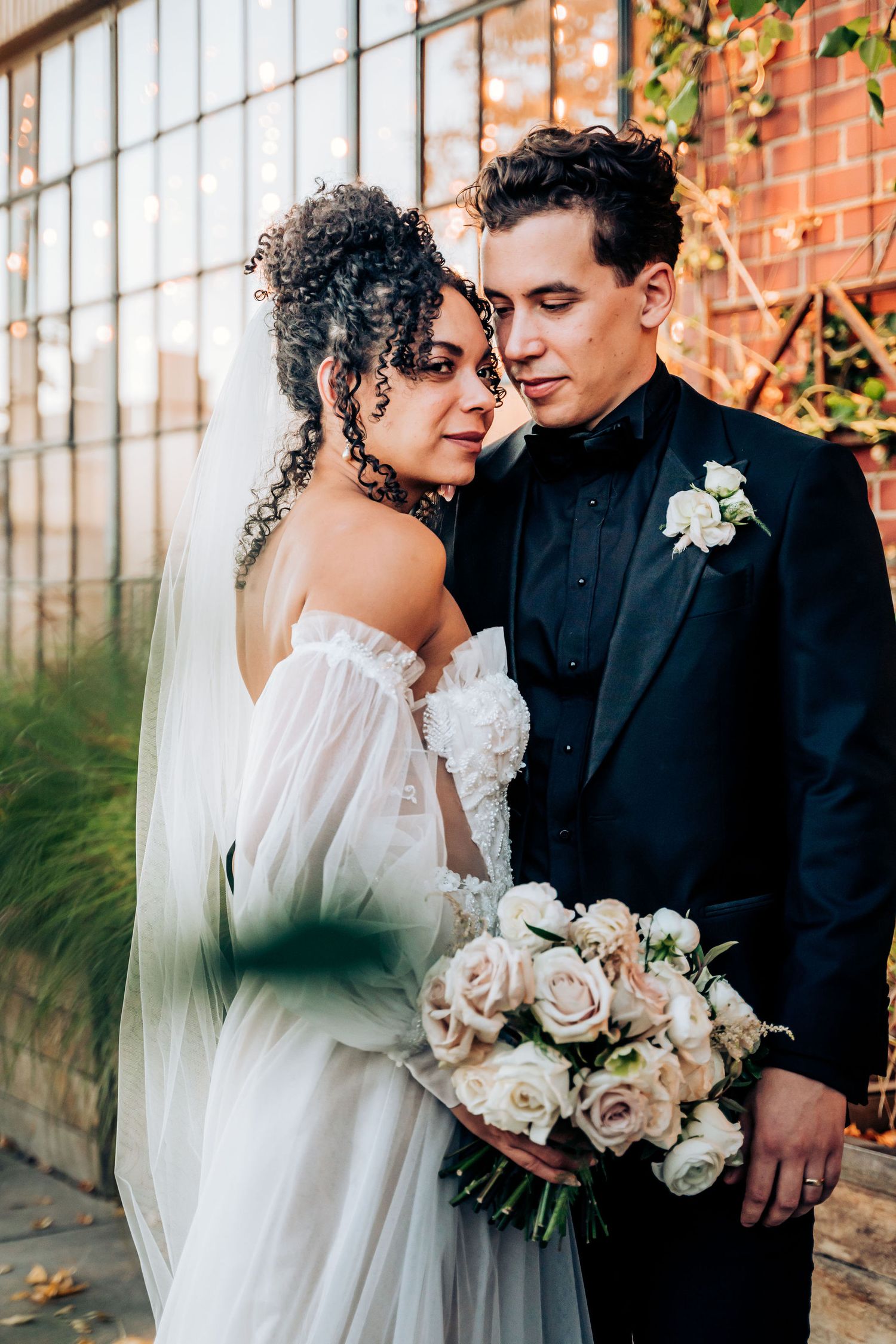 Bride wearing off-shoulder gown and groom in black suit share intimate moment with blush rose bouquet.