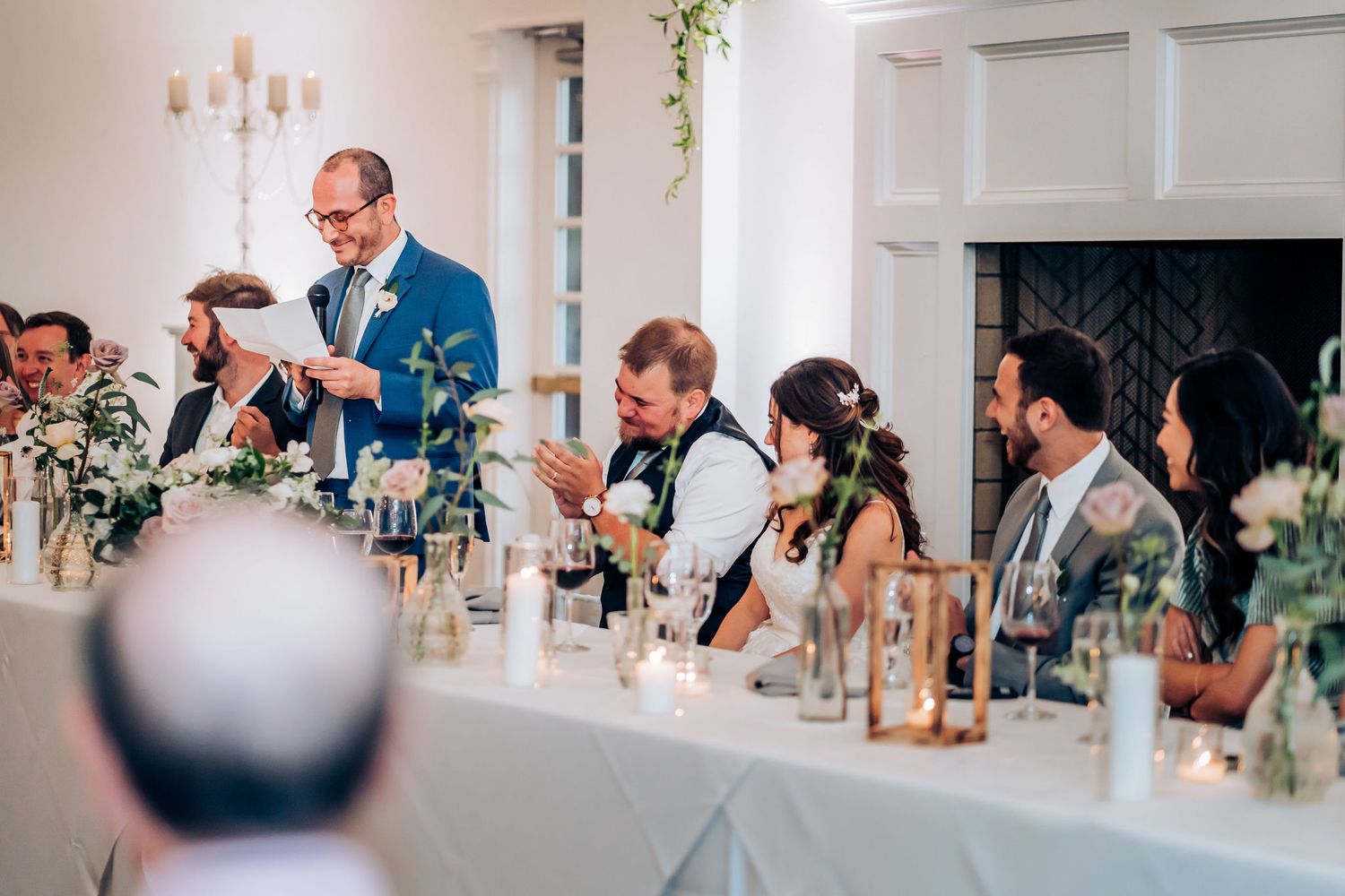 Wedding party members seated at head table listen to toasts during elegant reception.