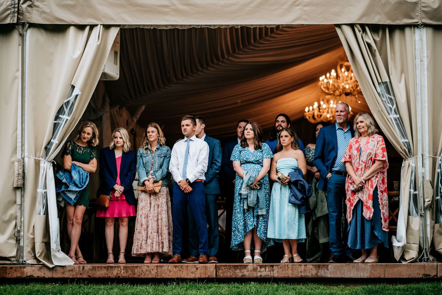 Wedding guests gather under an elegant tent decorated with chandeliers and draped fabric.