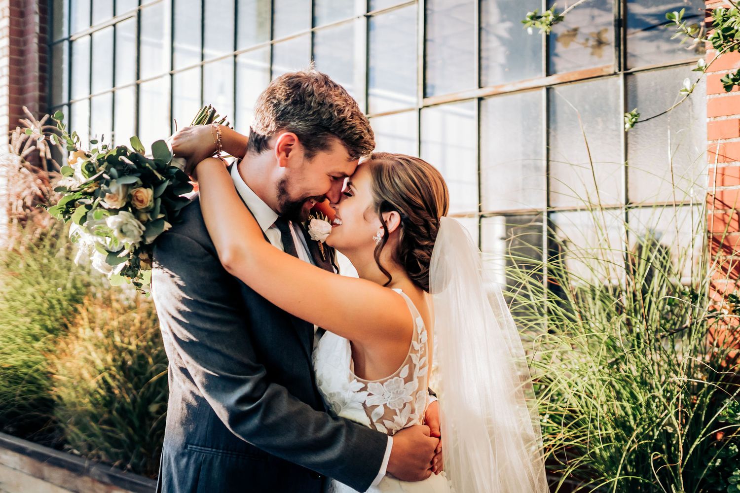 Romantic moment between newlyweds embracing outdoors near industrial windows and ornamental grasses.