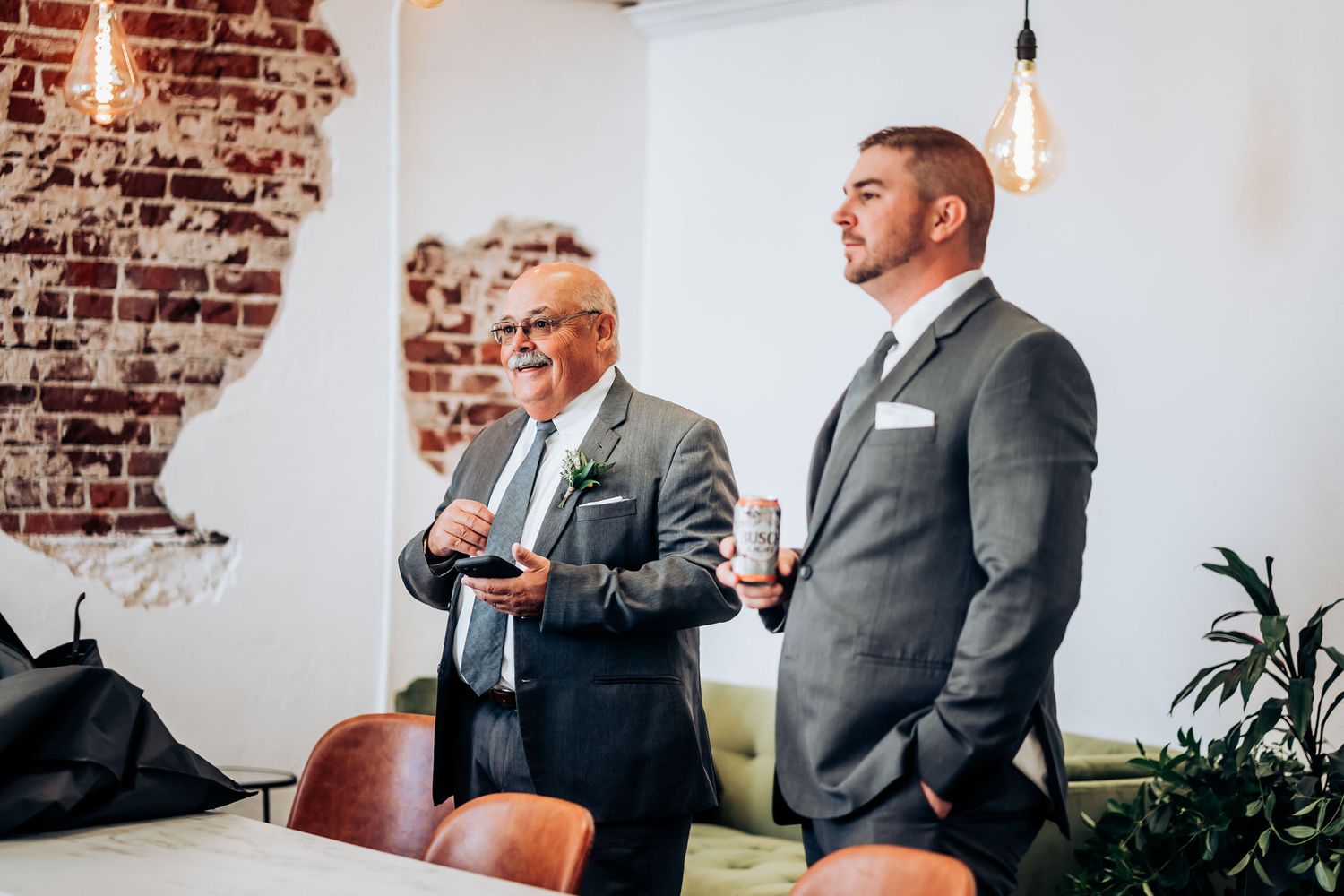 Two people in suits standing in a room with exposed brick walls and modern lighting fixtures.