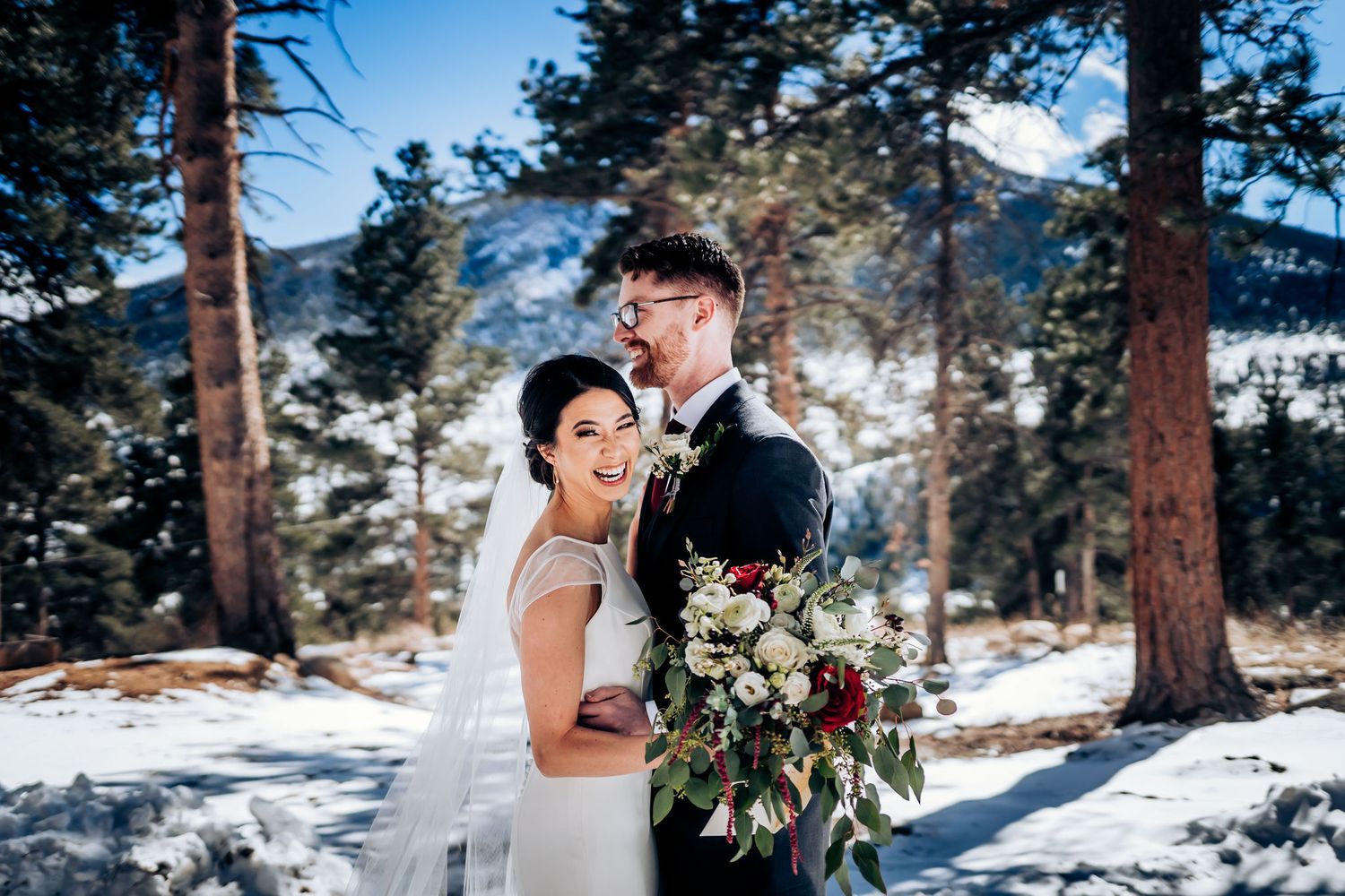 A romantic winter wedding portrait with the couple standing among snow-covered pine trees.