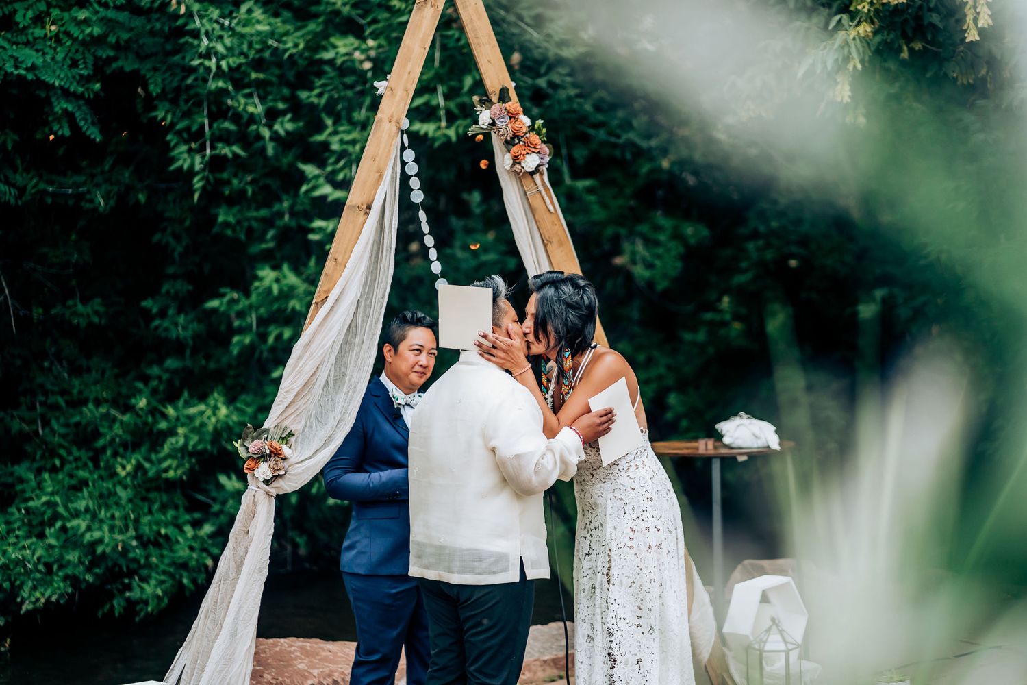 A wedding ceremony takes place under a wooden triangular arch decorated with white fabric in an outdoor setting.