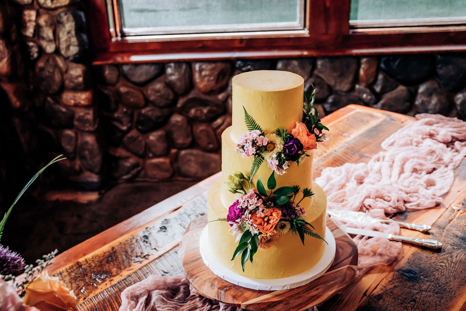 An elegant three-tiered wedding cake decorated with fresh flowers and greenery sits on a rustic wooden table.