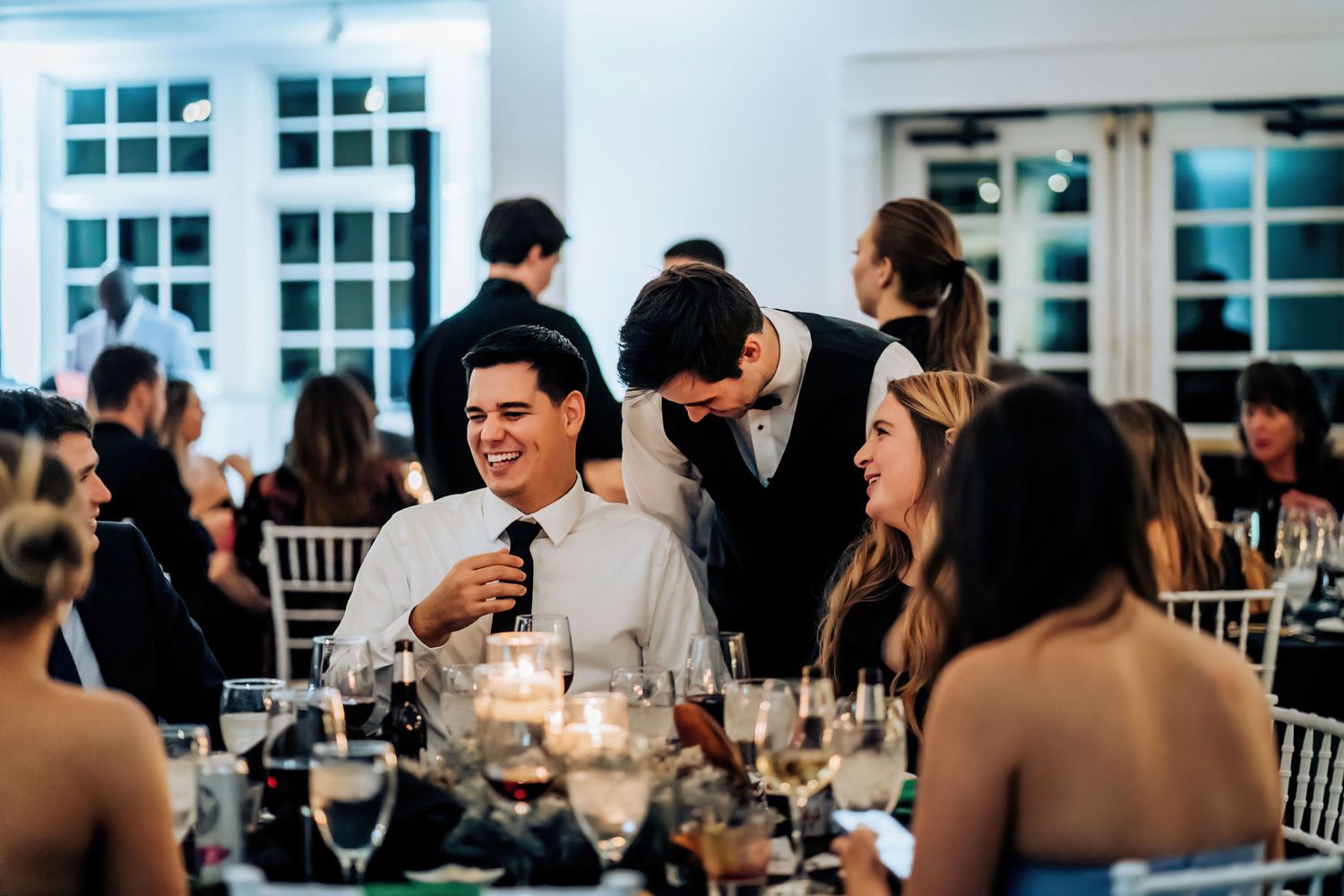 Wedding guests enjoying a lively dinner reception with elegant place settings and white decor.