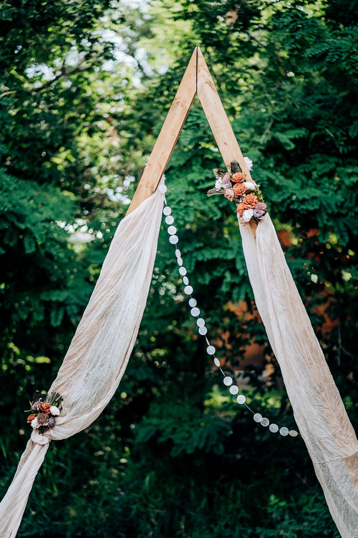Rustic triangular wedding arch with white fabric and floral accents against green foliage.