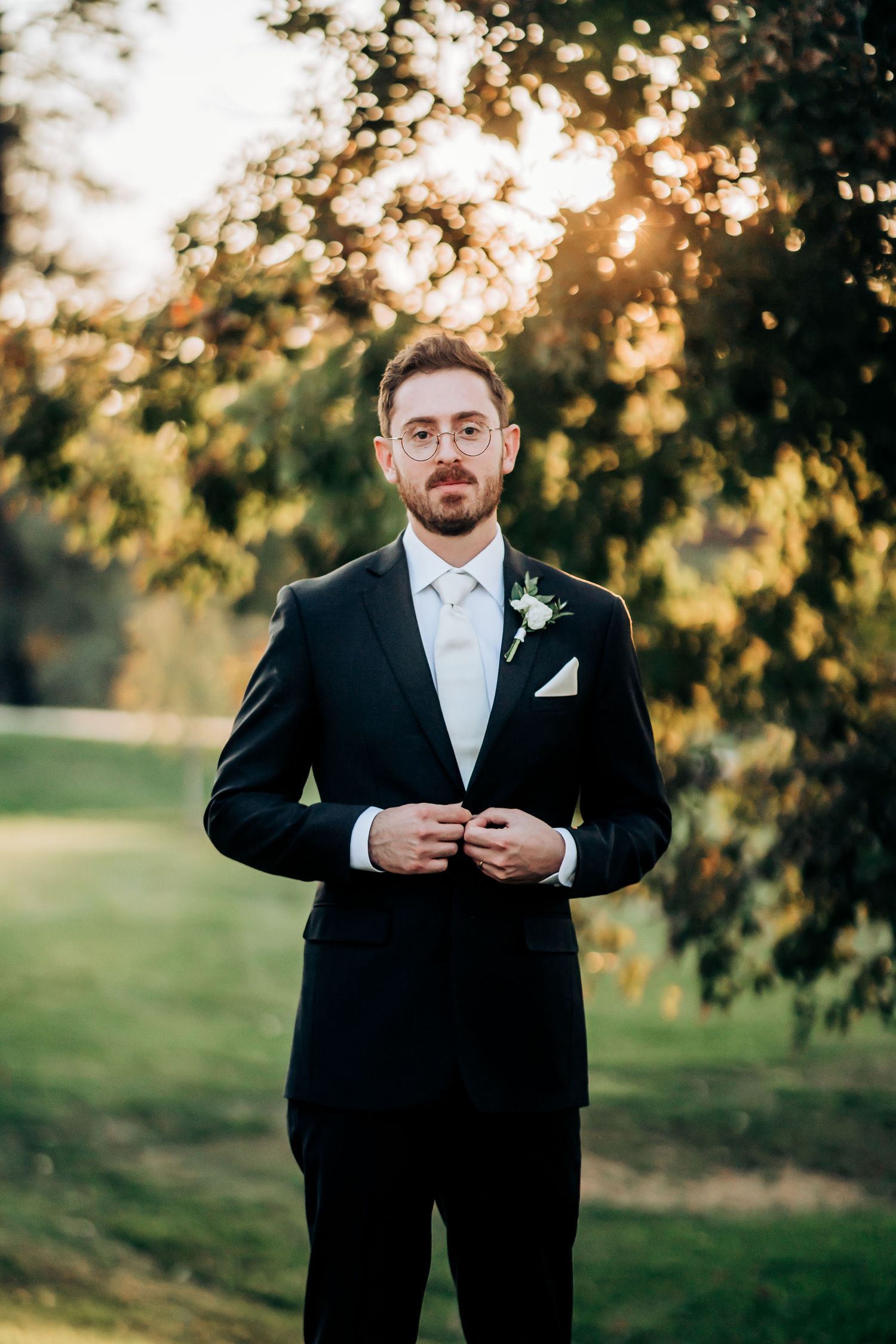 Formal portrait of groom in black tuxedo with boutonniere against sunset garden background.