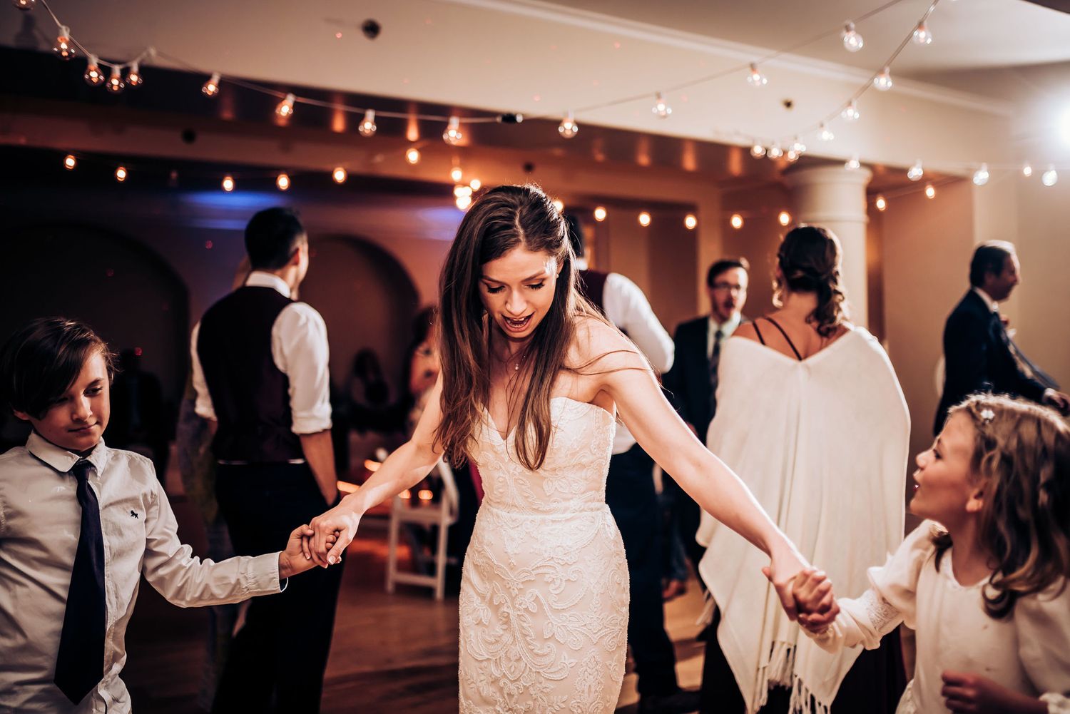 Dancing guests hold hands in a circle during a celebration in an elegant venue with string lights.