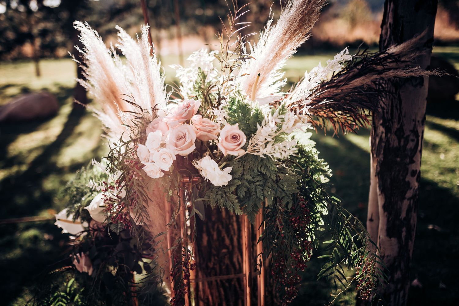 Romantic floral arrangement with pink roses and pampas grass against rustic wooden post.