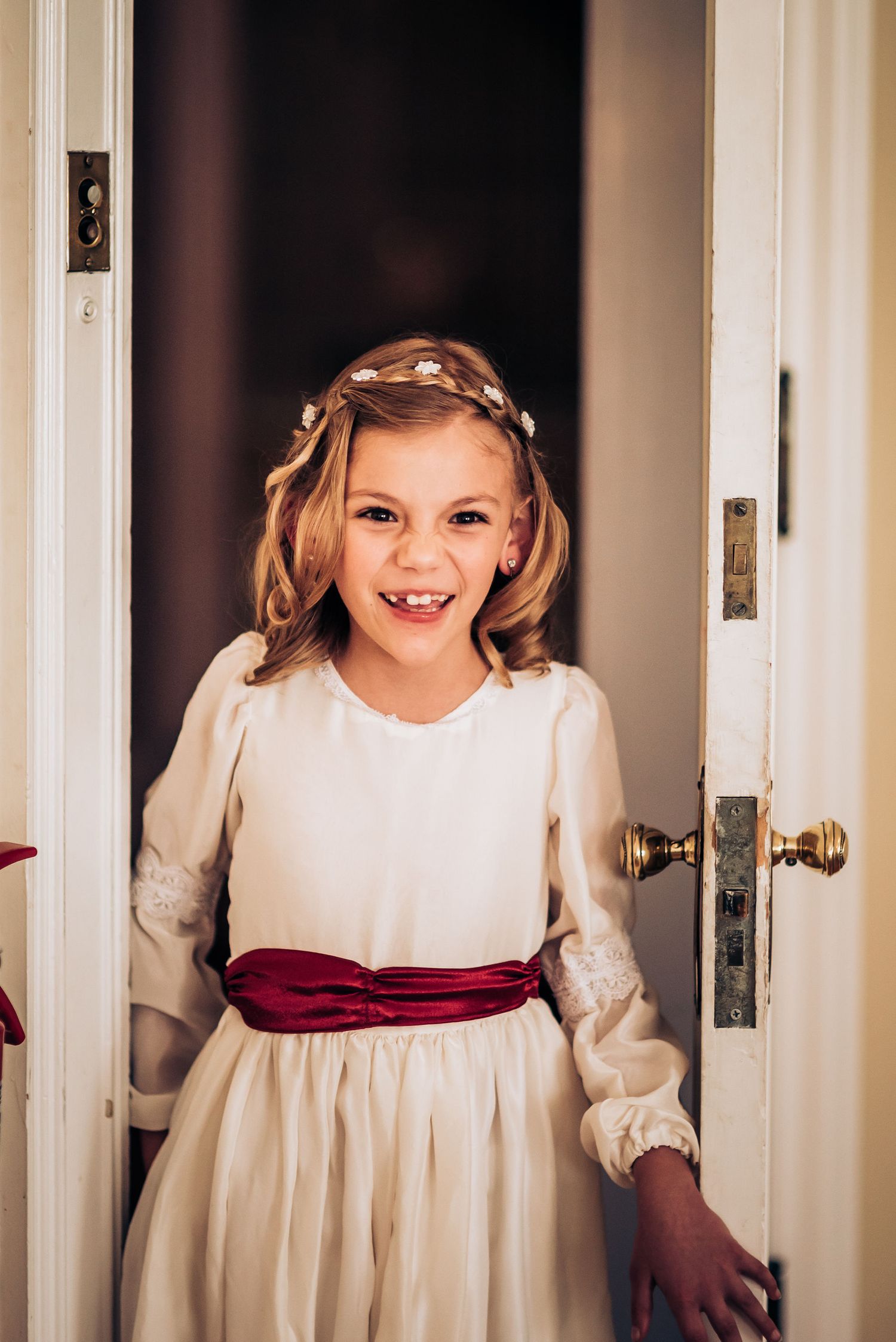 A young attendant in a cream-colored dress with burgundy sash stands in a doorway.