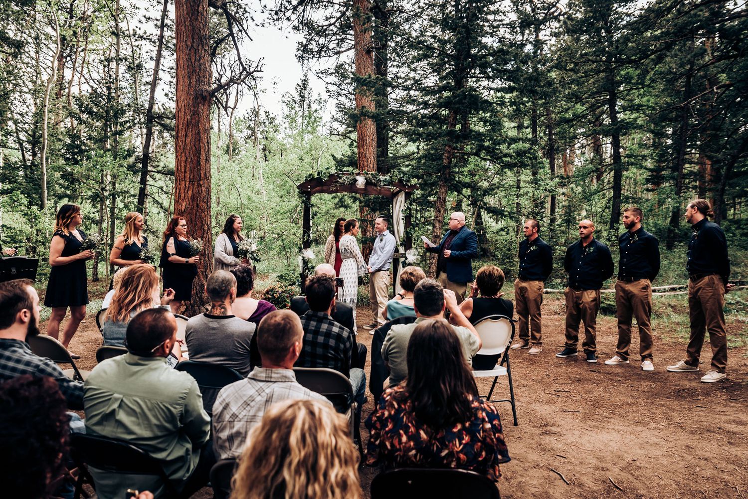 A forest wedding ceremony with guests seated on chairs and wedding party members standing in formation.