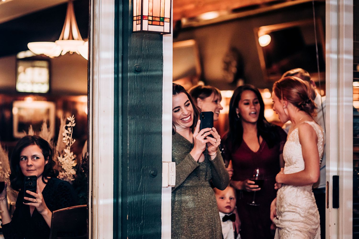 Wedding guests gather near glass doors taking photos and enjoying celebratory drinks.