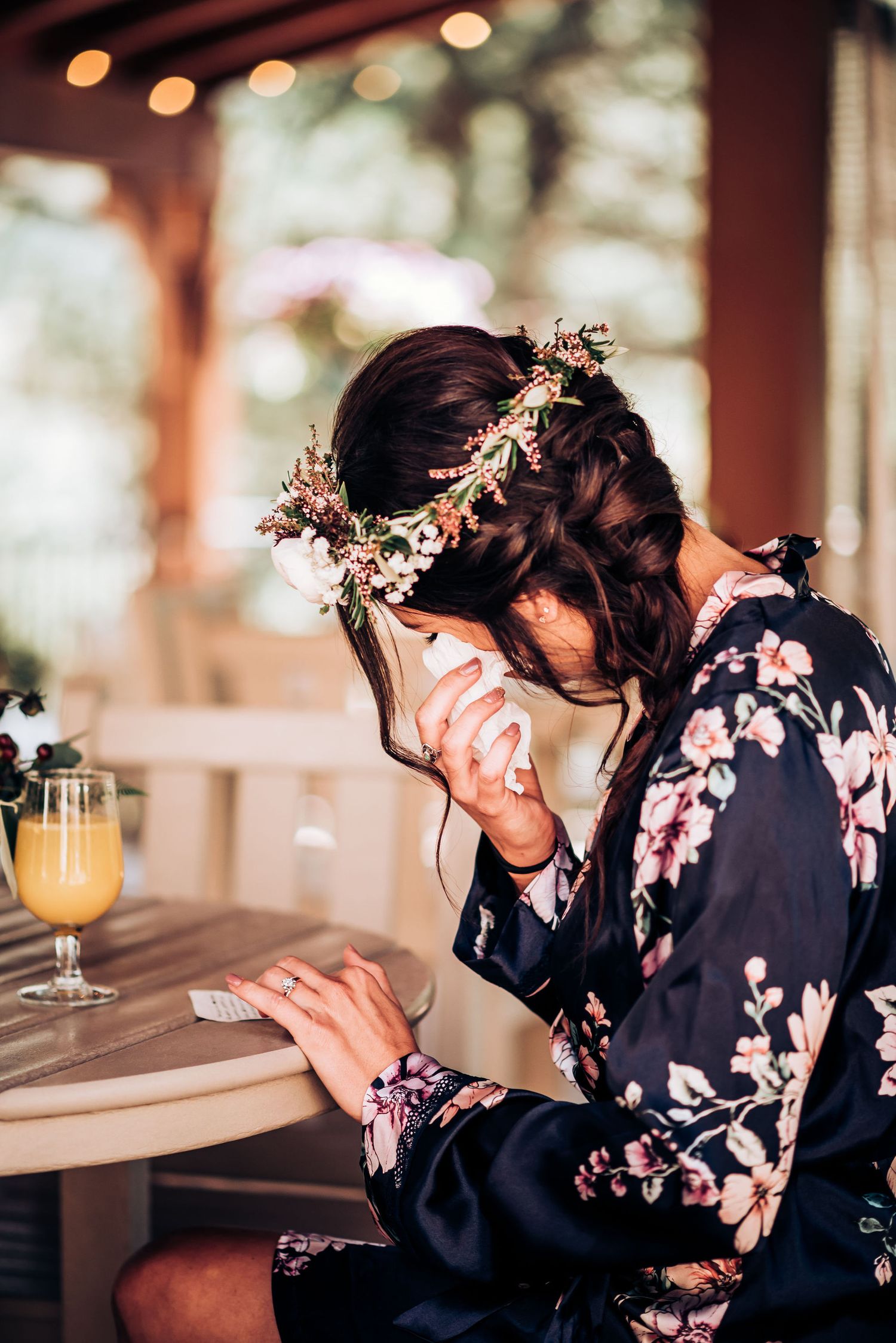 Delicate floral crown adorns a dark-haired figure in a floral robe sitting at a table with orange juice.