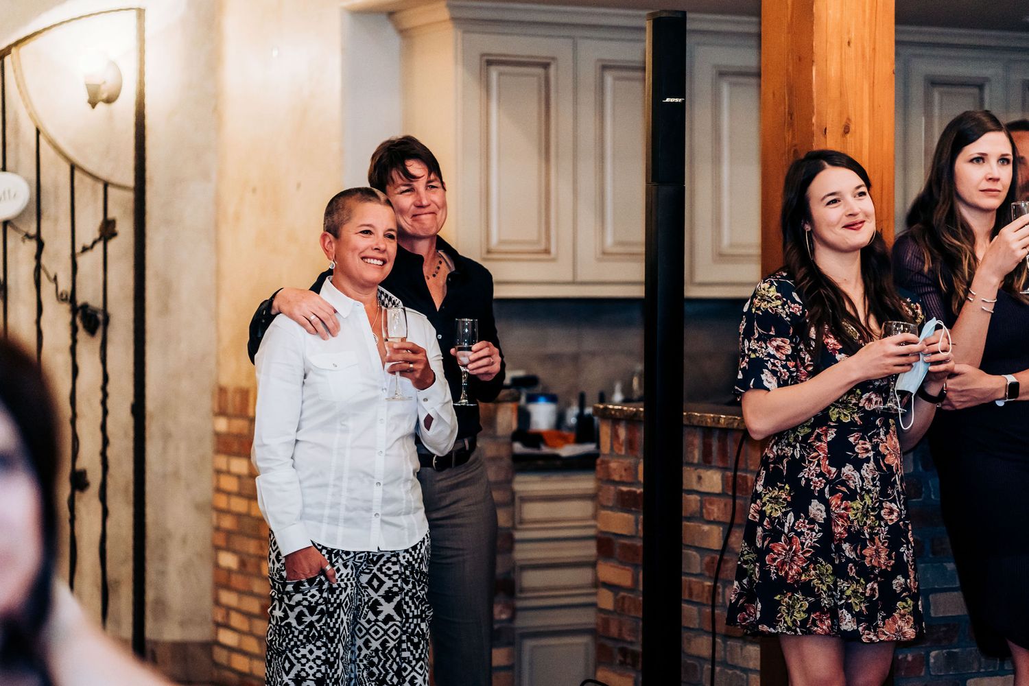 A group of people stand together at a social gathering, smiling and holding drinks in a rustic indoor setting.