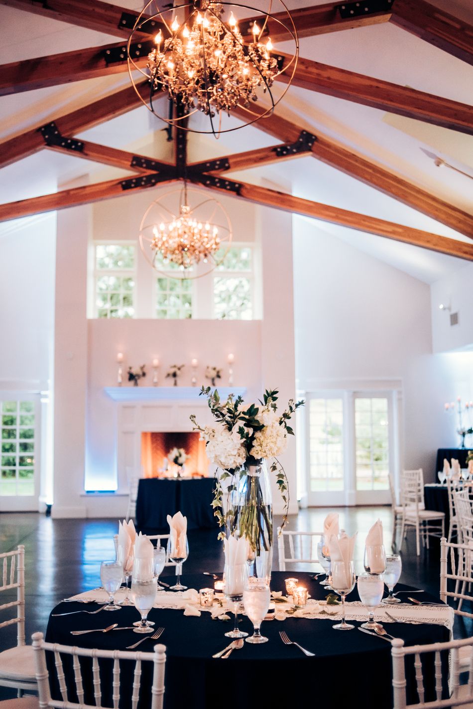 Elegant wedding reception table setting with crystal chandelier and exposed beam ceiling in white venue space.