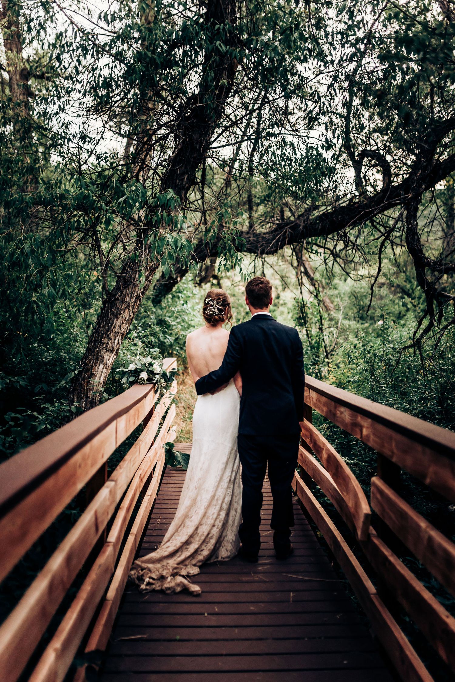 Newlyweds stand together on wooden bridge surrounded by lush forest canopy.