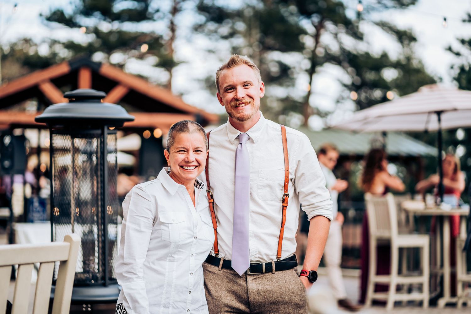 Two people in formal attire stand together on an outdoor patio with pine trees in the background.