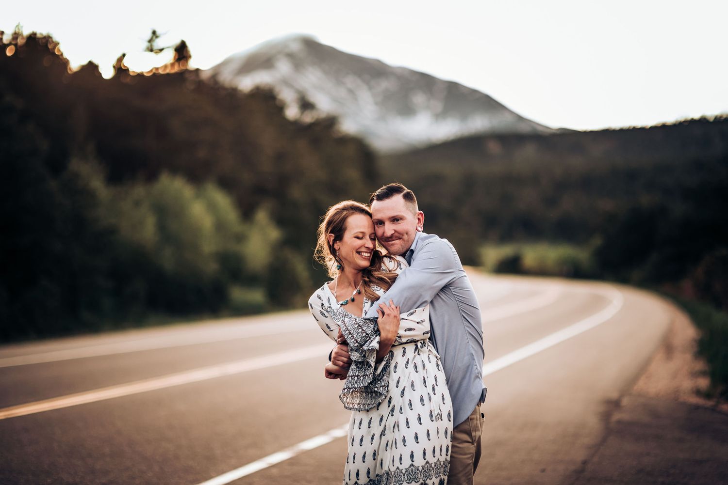 Joyful couple embracing on mountain road with snow-capped peaks in distance.